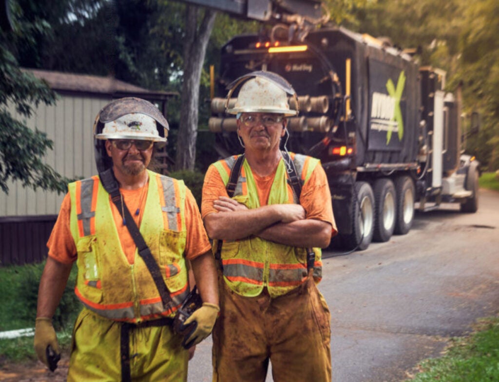 two men in work gear standing in from of a hydro x truck